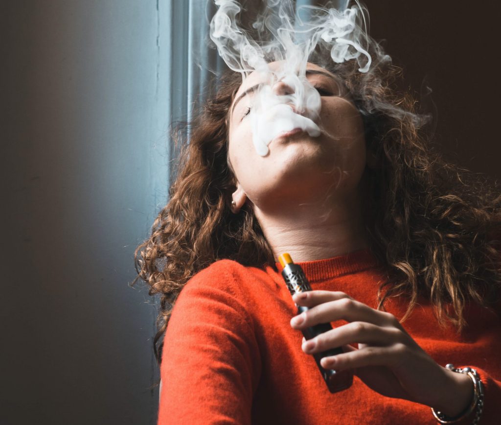 femme avec e-cigarette exhalant de la vapeur blanche, portant un pull orange