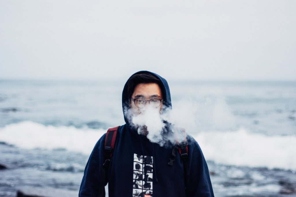 jeune homme avec lunettes vaporisant devant la mer, concept produit tabac chauffé