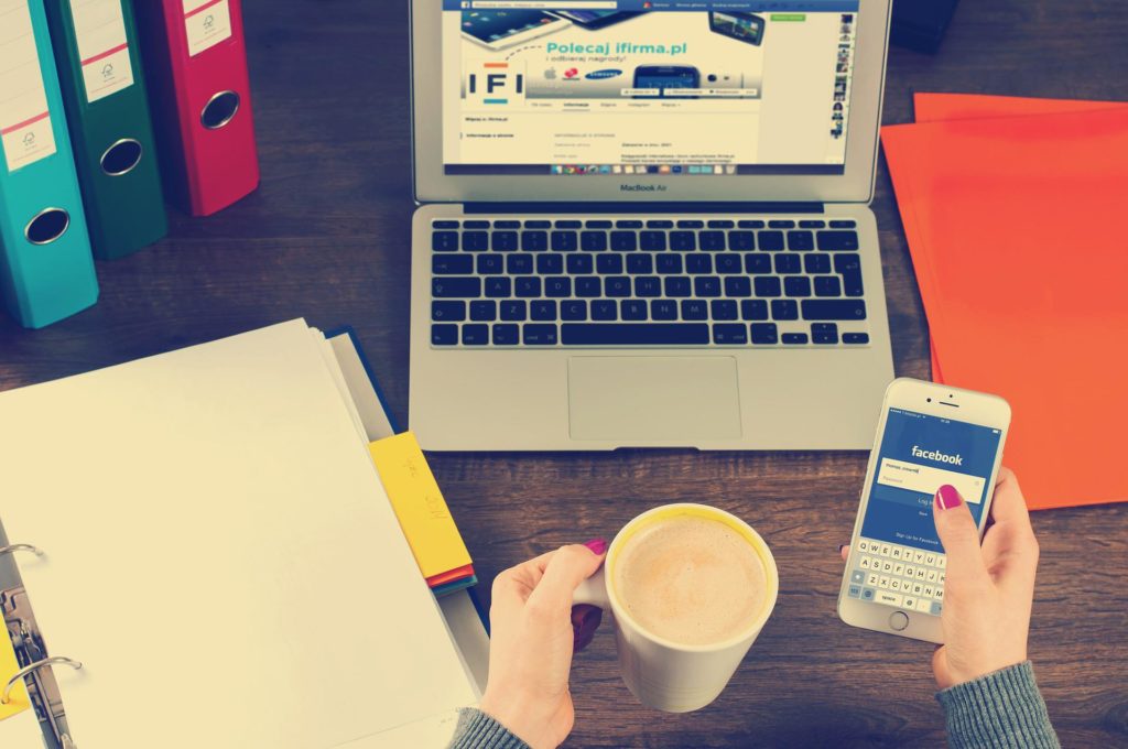 une personne tient un café et consulte facebook sur son smartphone à son bureau avec un laptop