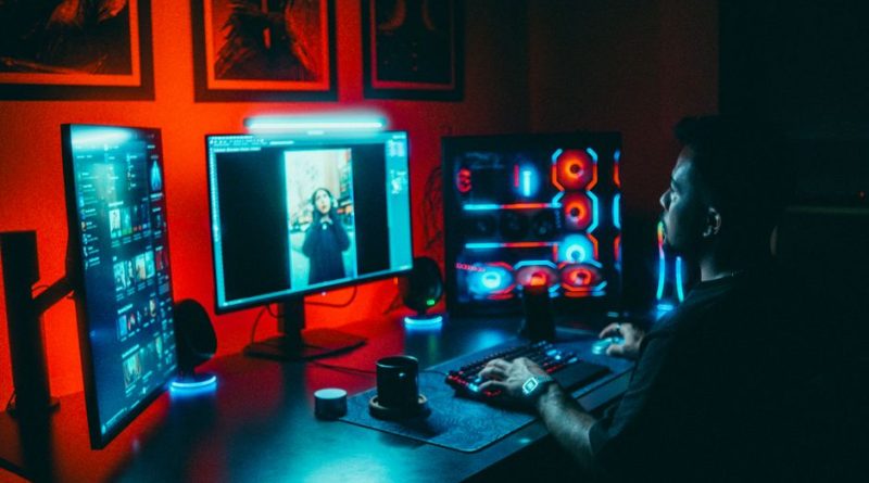 A young man working on a multi-screen computer setup in a dark, neon-lit room.